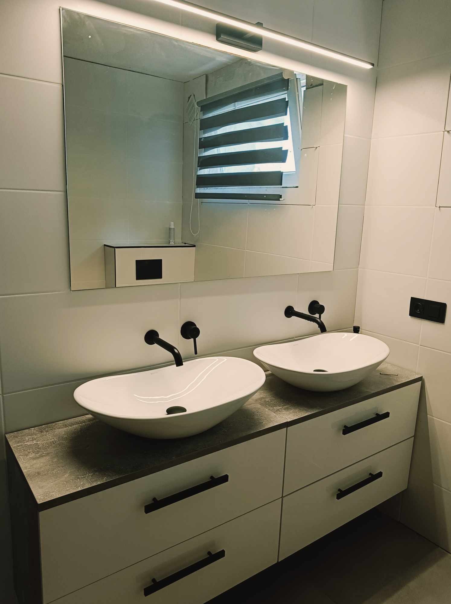 Nowoczesna łazienka z dwiema owalnymi umywalkami nablatowymi i czarną armaturą. Białe płytki na ścianach, lustro z podświetleniem LED i szafka z szufladami.