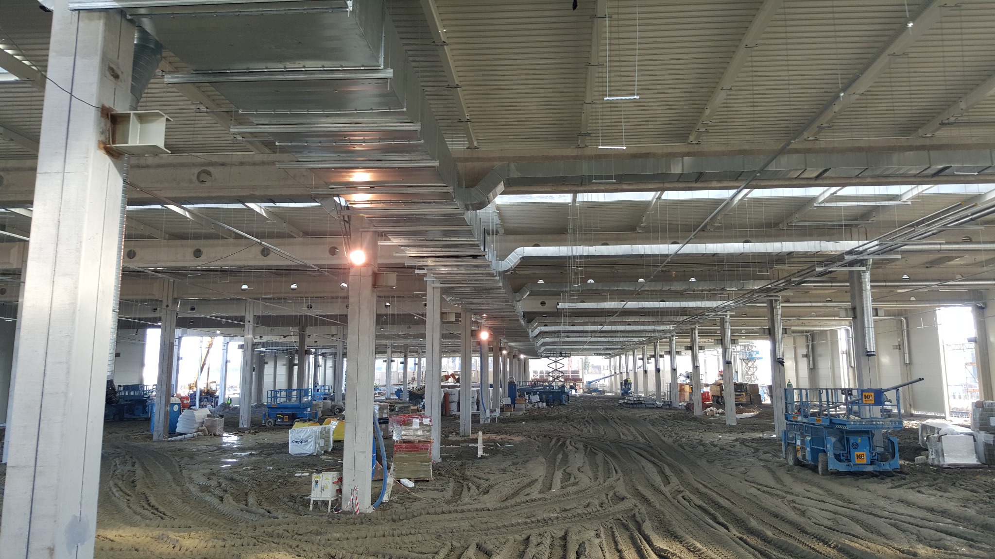 Rozległa hala w trakcie montażu wentylacji: widoczne kanały wentylacyjne pod sufitem, betonowe kolumny i maszyny budowlane na ziemi. Oświetlenie robocze.