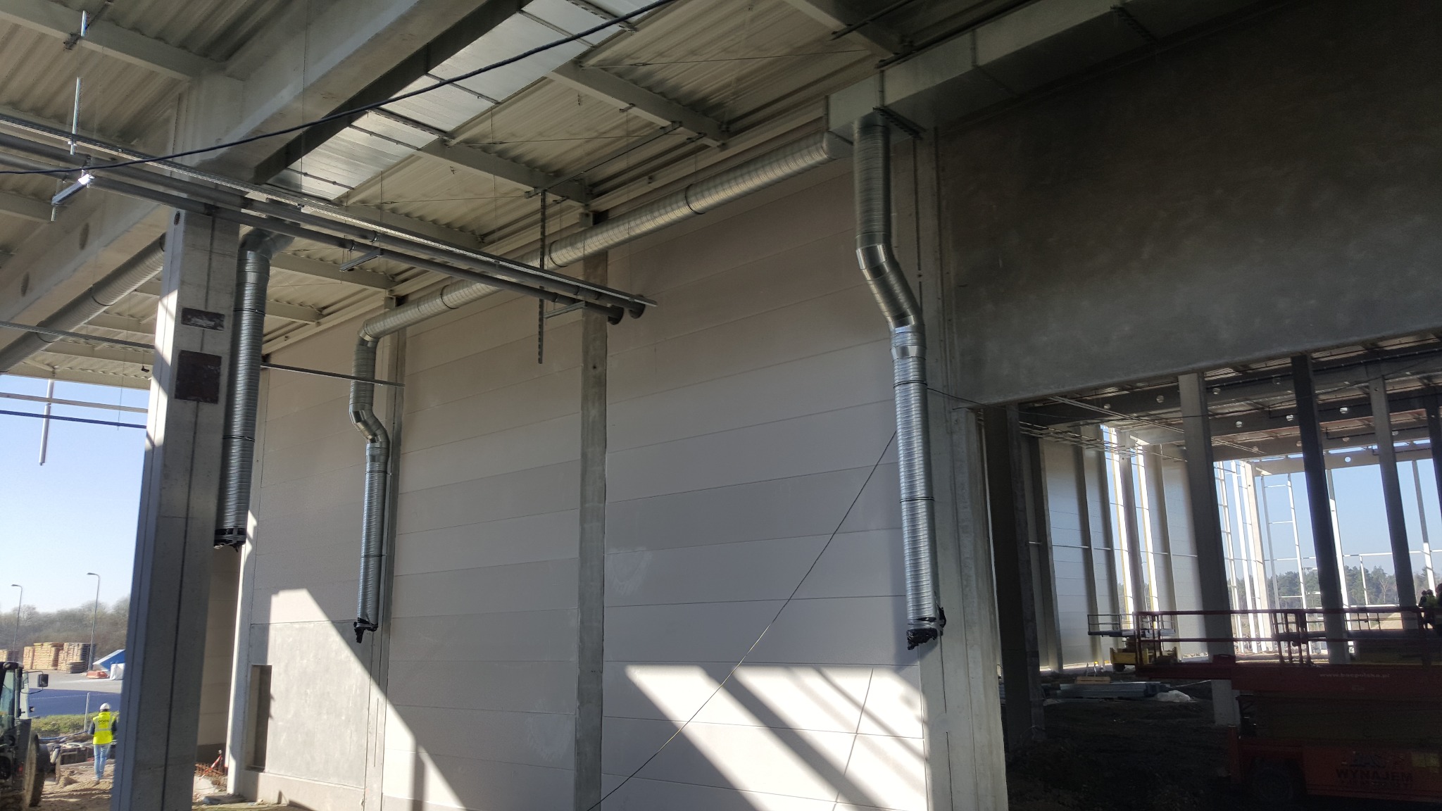 Wnętrze hali w budowie z zamontowanymi, spiralnymi przewodami wentylacyjnymi przy ścianach i pod sufitem. Widoczny surowy beton i konstrukcja budynku.