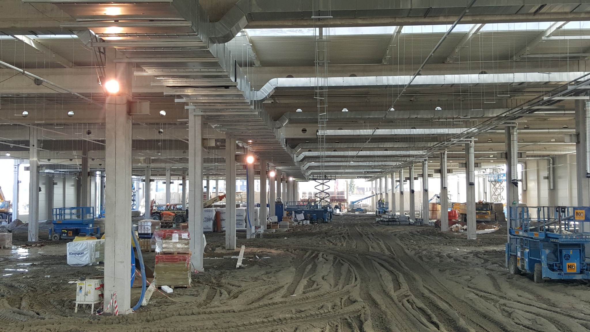 Wnętrze hali w trakcie montażu instalacji wentylacyjnej. Widoczne kanały wentylacyjne pod sufitem, kolumny nośne i sprzęt budowlany na ziemi.