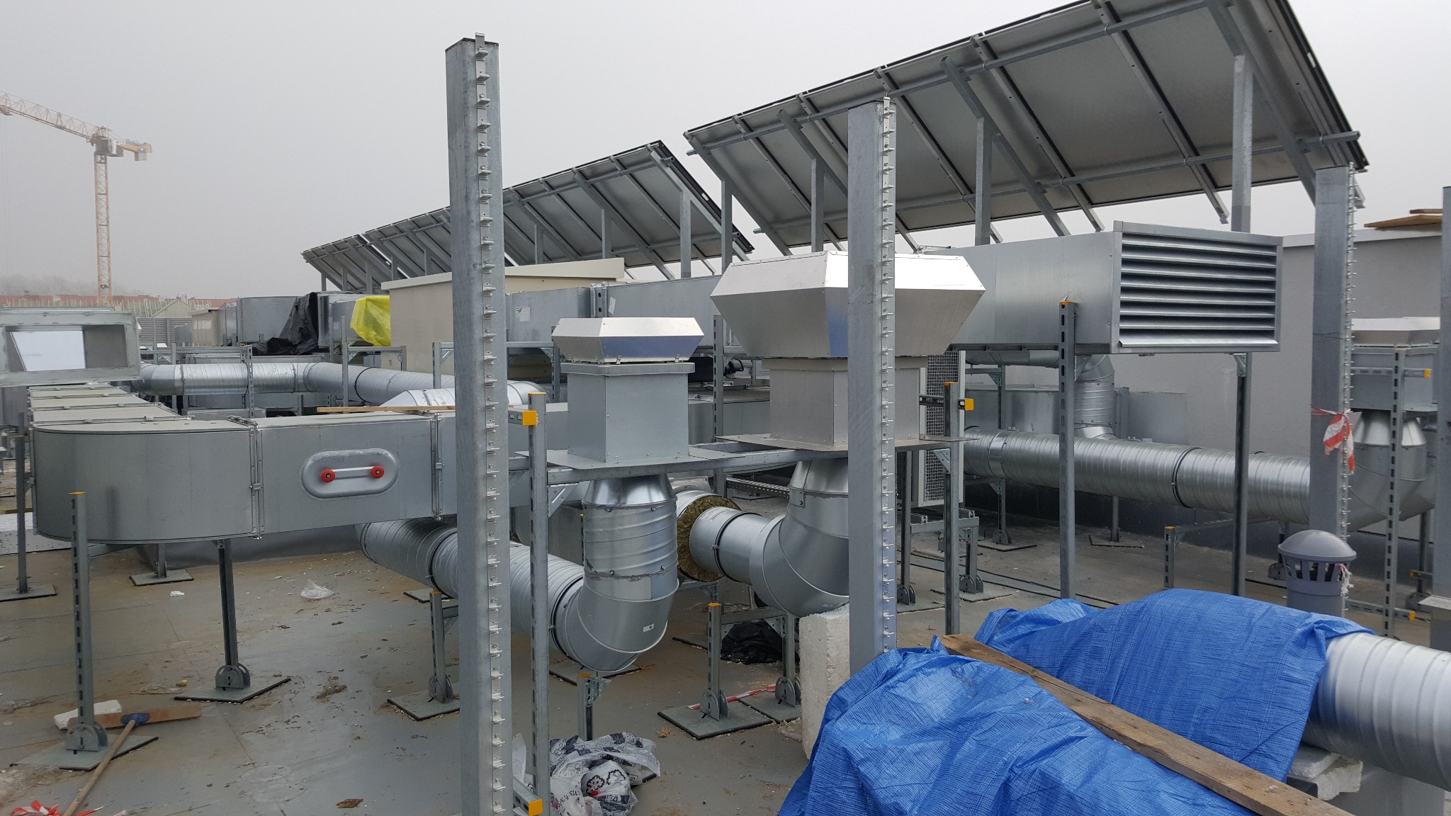 Kompleksowy system wentylacyjny na dachu budynku komercyjnego, z widocznymi kanałami, centralami i elementami konstrukcyjnymi. W tle dźwig budowlany.