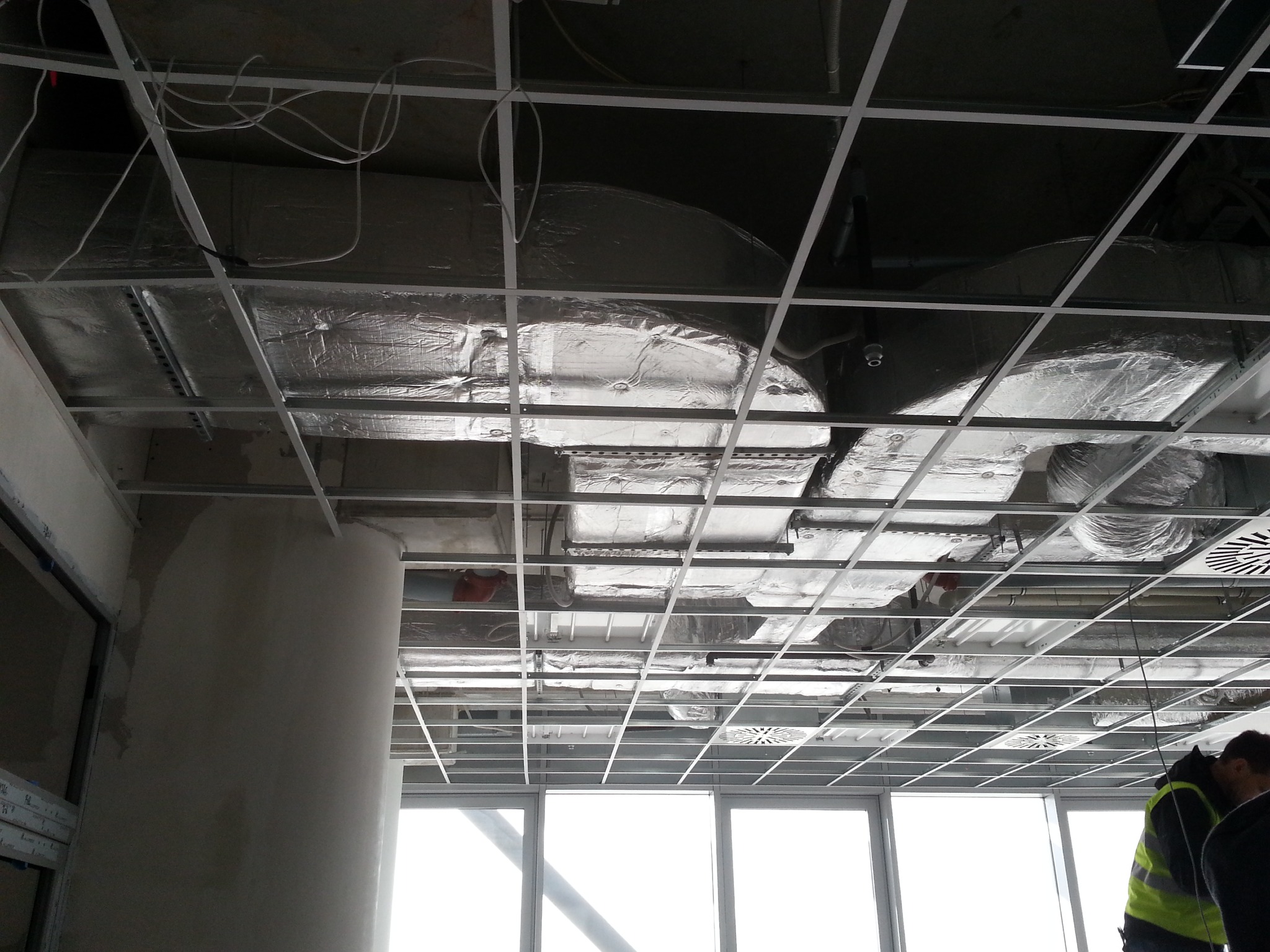 Montaż instalacji wentylacyjnej w budynku komercyjnym. Widoczne srebrne rury wentylacyjne pod sufitem, metalowa konstrukcja podwieszana i fragment okna.