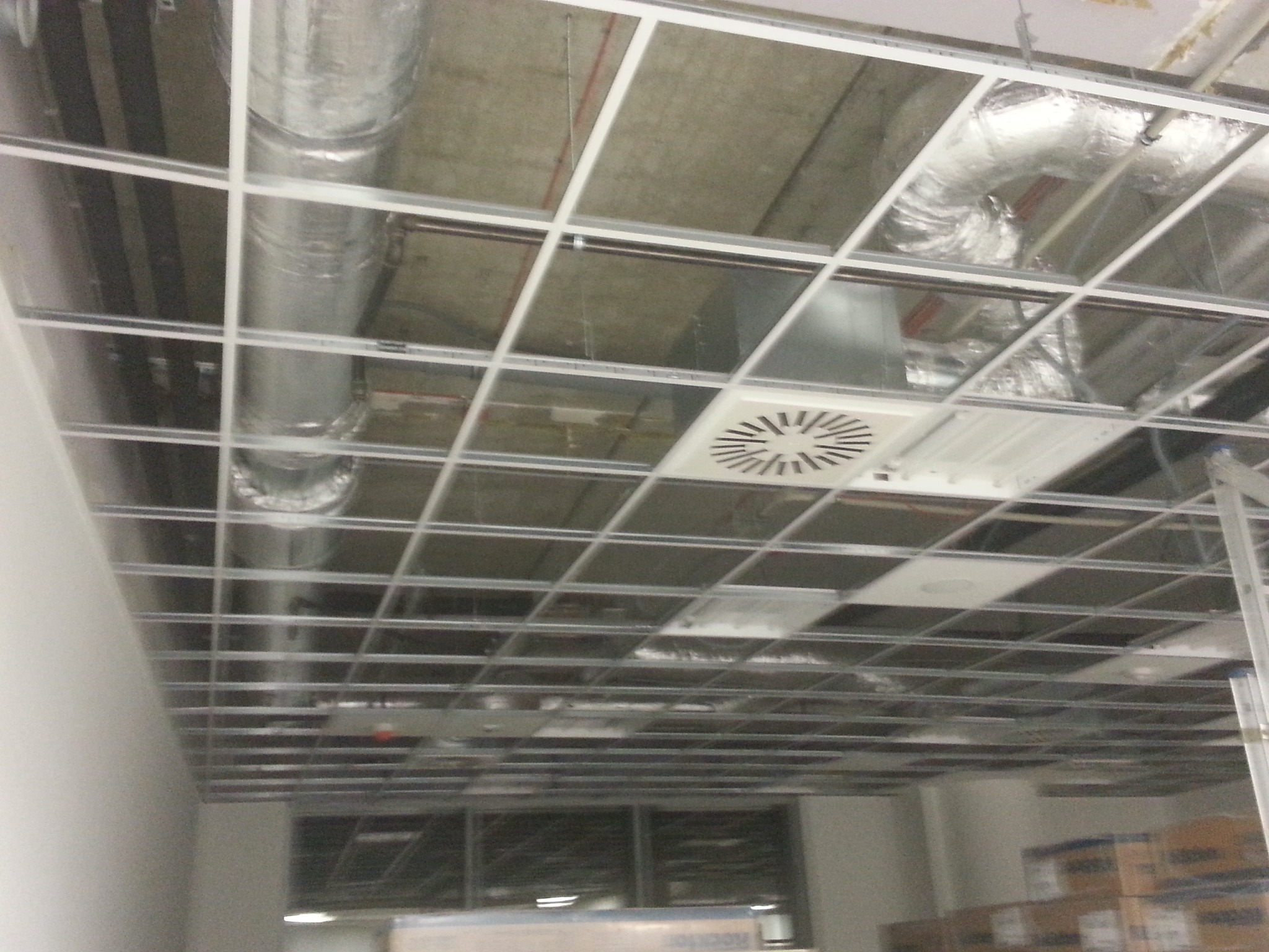 Sufit podwieszany z widocznymi elementami instalacji wentylacyjnej, w tym okrągłymi kanałami i kratką wentylacyjną. Widok z dołu, surowy beton w tle.