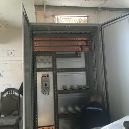 Mariusz Janczewski - Otwarta szafa elektryczna z widocznymi elementami instalacji, miedzianymi szynami i zabezpieczeniami. W tle fragment pomieszczenia z płytkami i krzesłem.