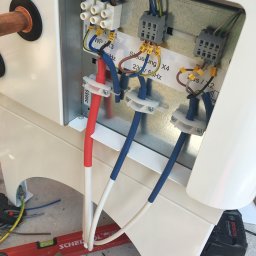 Mariusz Janczewski - Podłączenie elektryczne pieca gazowego w Warszawie. Widoczne kable w kolorach czerwonym i niebieskim, złączki oraz narzędzia pracy na podłodze.