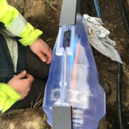 Mariusz Janczewski - Elektryk łączy kable w ziemi za pomocą specjalnej mufy. Widoczne ręce w odblaskowej kurtce i fragment kabli w wykopie. Prace instalacyjne przyłącza elektrycznego.