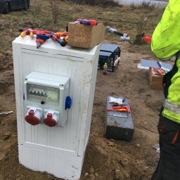 Mariusz Janczewski - Biała skrzynka elektryczna z gniazdami i bezpiecznikami, narzędzia na górze i obok, ziemiste tło z jeziorem w oddali, częściowo widoczna osoba w odblaskowej kurtce.