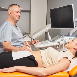 AB Clinic Centrum Ginekologiczno-Położnicze - Lekarz uśmiechnięty wykonuje USG jamy brzusznej pacjentce leżącej na pomarańczowym leżaku w gabinecie z nowoczesną aparaturą.