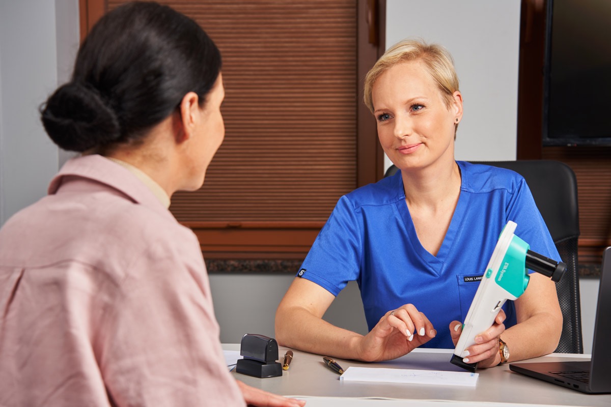 Ginekolog podczas konsultacji z pacjentką, trzyma urządzenie diagnostyczne EVA System. Na biurku dokumenty, pieczątka i laptop. Jasne wnętrze gabinetu.