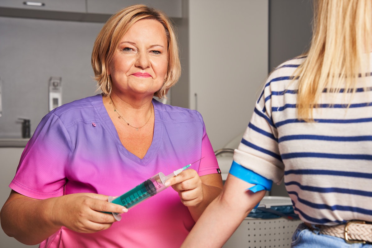 Lekarka w fioletowo-różowym uniformie trzyma strzykawkę z zielonym płynem, przygotowując się do pobrania krwi od pacjentki. Widoczny niebieski pasek na ramieniu.