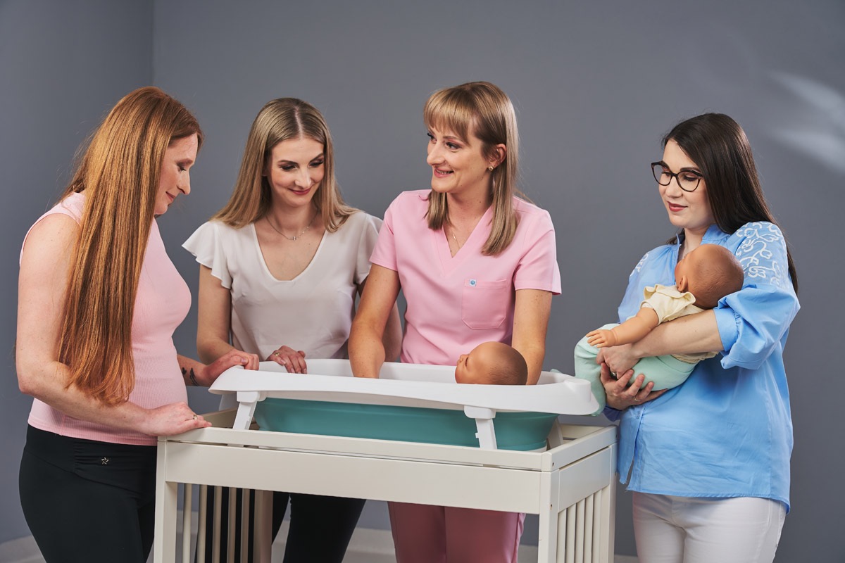 Personel medyczny demonstruje kąpiel noworodka na specjalnym przewijaku z wanienką, edukując przyszłe mamy w szpitalu. Jasne, stonowane kolory.