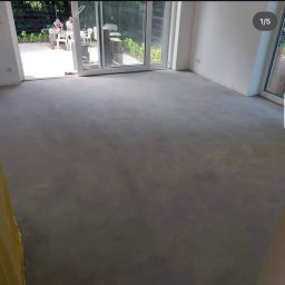 Cezary Stradowski - Jasnoszara, jednolita posadzka w minimalistycznym wnętrzu z dużymi oknami tarasowymi, przez które widać osoby siedzące w ogrodzie.