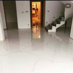 Cezary Stradowski - Błyszcząca, biała posadzka z subtelnym marmurkowym wzorem w nowym wnętrzu. Widoczne schody w tle i otwarte przejście do kolejnego pomieszczenia.