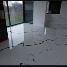 Biała, błyszcząca posadzka z czarnymi żyłkami w nowoczesnym wnętrzu z dużym oknem. Widoczne odbicia światła podkreślają gładką powierzchnię podłogi.