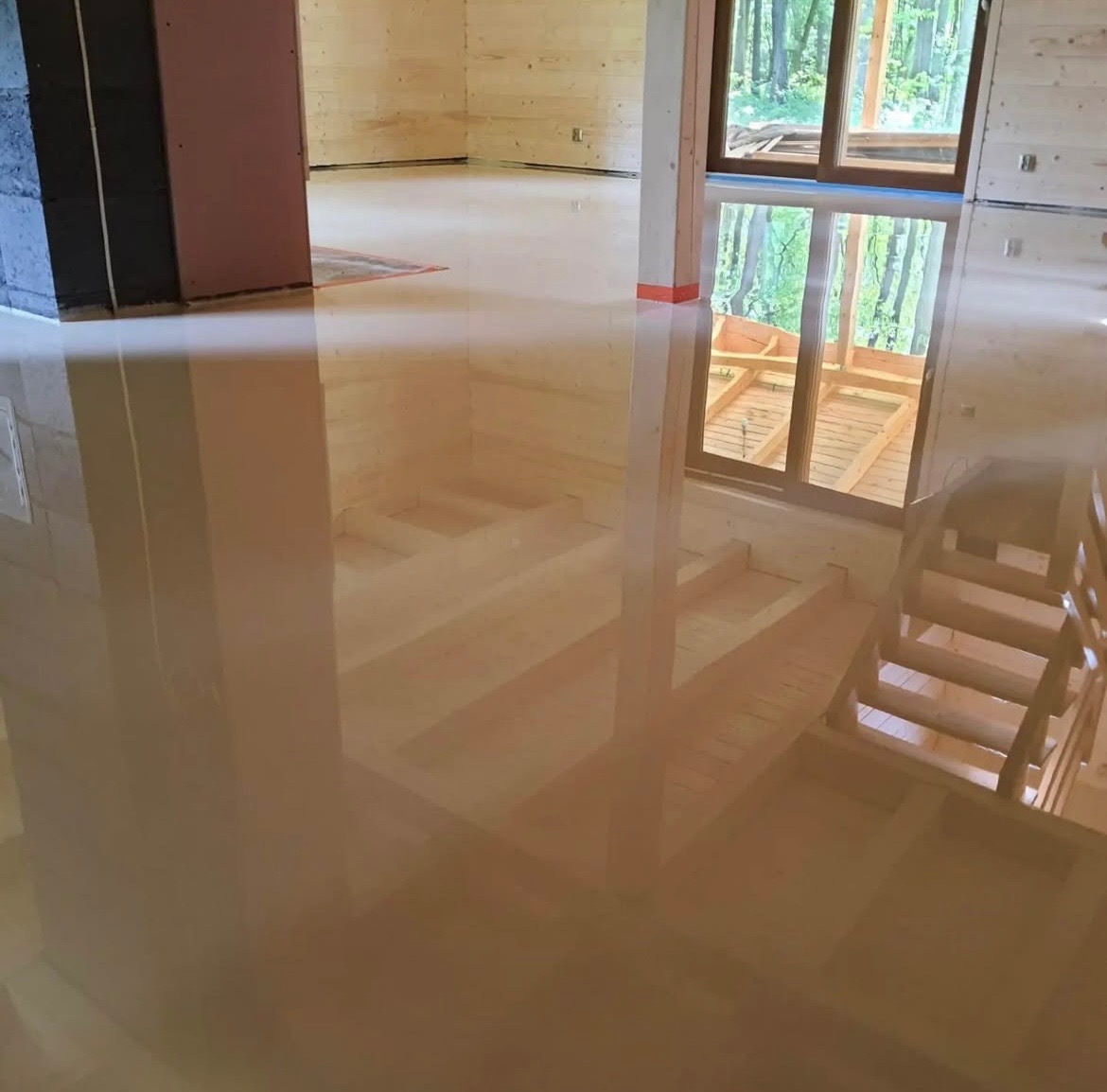 Błyszcząca, beżowa posadzka w surowym wnętrzu drewnianego domu, z widocznym odbiciem okna i schodów. Minimalistyczny design, gładka powierzchnia.