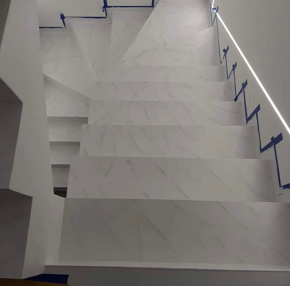 Białe schody z marmurkowym wzorem, zabezpieczone niebieską taśmą malarską. Oświetlenie LED wzdłuż ściany. Widok z góry na wykończone schody.