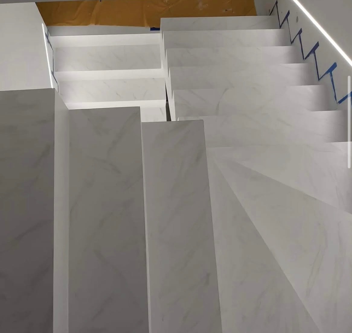 Białe schody wykończone płytami imitującymi marmur, widok z góry, oświetlenie LED wzdłuż krawędzi, zabezpieczone taśmą malarską, nowoczesny design.