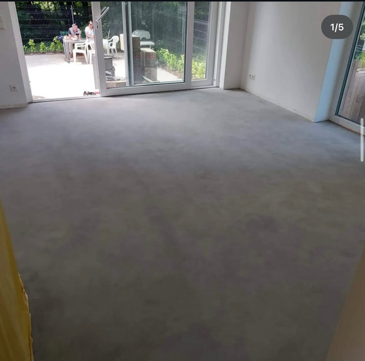 Jasnoszara, jednolita posadzka w minimalistycznym wnętrzu z dużymi oknami tarasowymi, przez które widać osoby siedzące w ogrodzie.