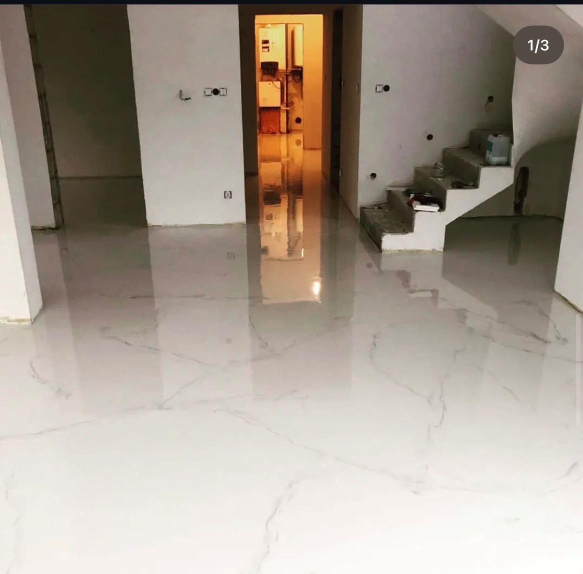 Błyszcząca, biała posadzka z subtelnym marmurkowym wzorem w nowym wnętrzu. Widoczne schody w tle i otwarte przejście do kolejnego pomieszczenia.