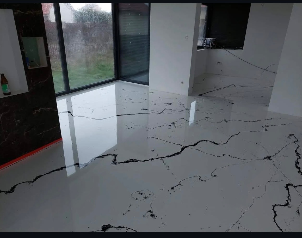 Biała, błyszcząca posadzka z czarnymi żyłkami w nowoczesnym wnętrzu z dużym oknem. Widoczne odbicia światła podkreślają gładką powierzchnię podłogi.