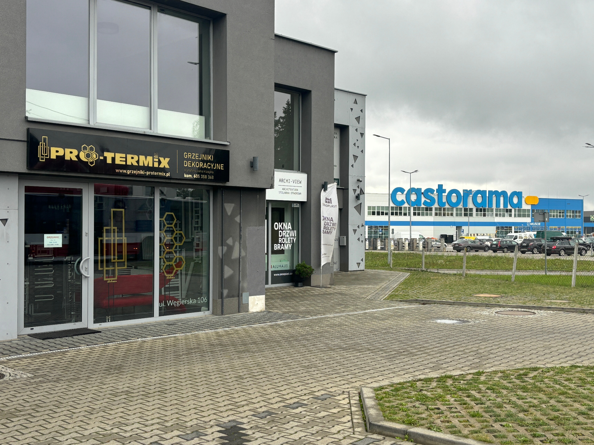 Fasada budynku firmy Pro-Termix z grzejnikami dekoracyjnymi i oknami, na tle Castoramy. Widoczny szyld z nazwą firmy i adresem ul. Węgierska 106.