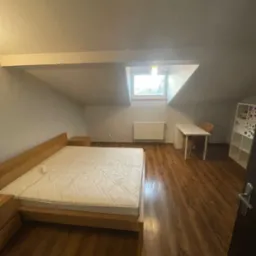 Pokój na poddaszu z drewnianą podłogą, łóżkiem i oknem dachowym. Ściany pomalowane na jasny kolor. Widoczne biurko, krzesło i regał.