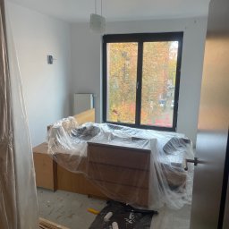 ANATOLII FILIPOV GOLD TOUCH - Pokój w trakcie malowania: meble zabezpieczone folią, wałki malarskie na podłodze, widok na jesienne drzewa za oknem. Świeżo pomalowane ściany w jasnym odcieniu.