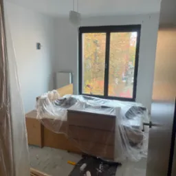 Pokój w trakcie malowania: meble zabezpieczone folią, wałki malarskie na podłodze, widok na jesienne drzewa za oknem. Świeżo pomalowane ściany w jasnym odcieniu.