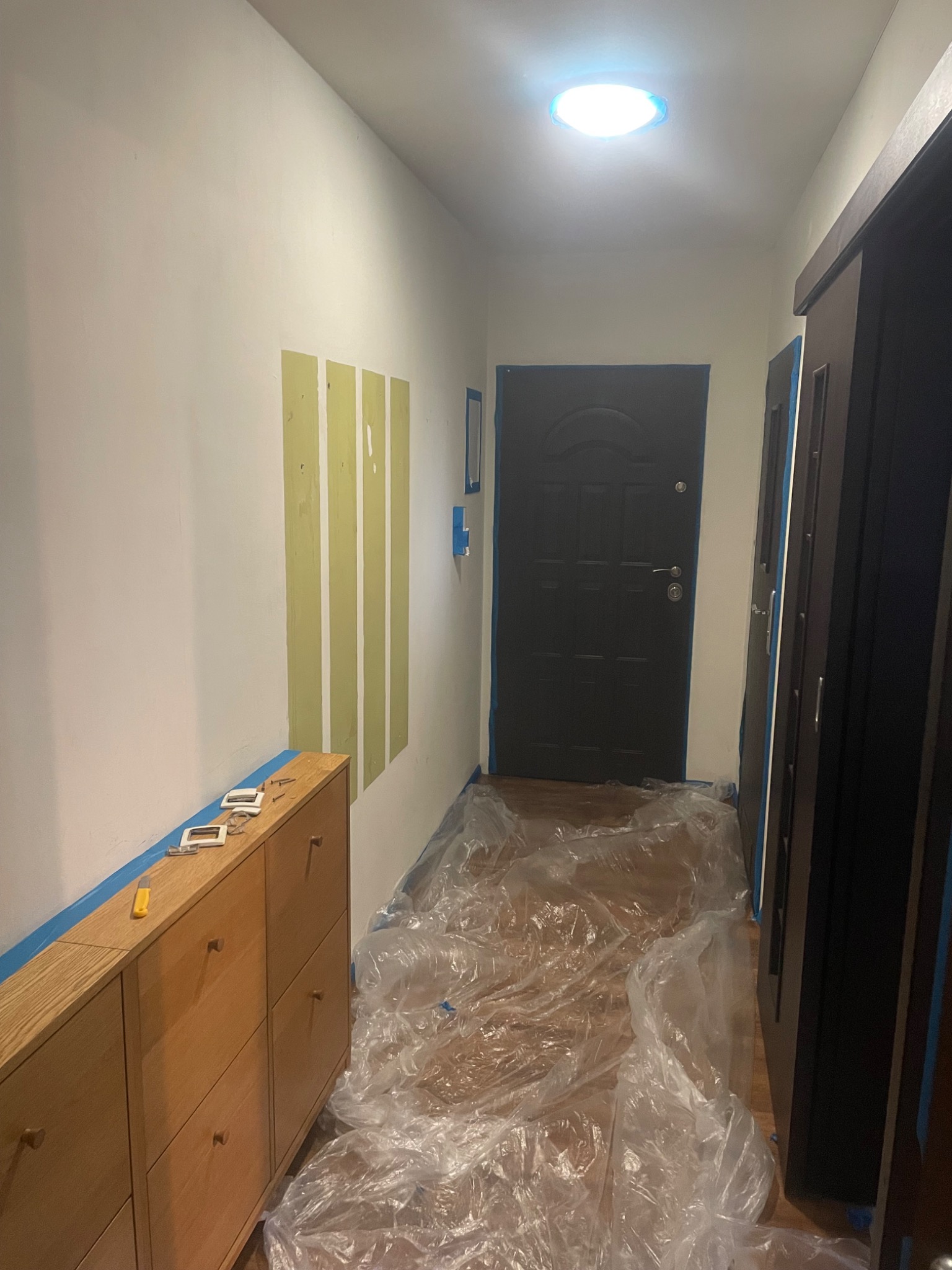 Korytarz w trakcie malowania: ściany z fragmentami zagruntowanymi pod farbę, meble zabezpieczone taśmą, podłoga przykryta folią ochronną, widoczne narzędzia.