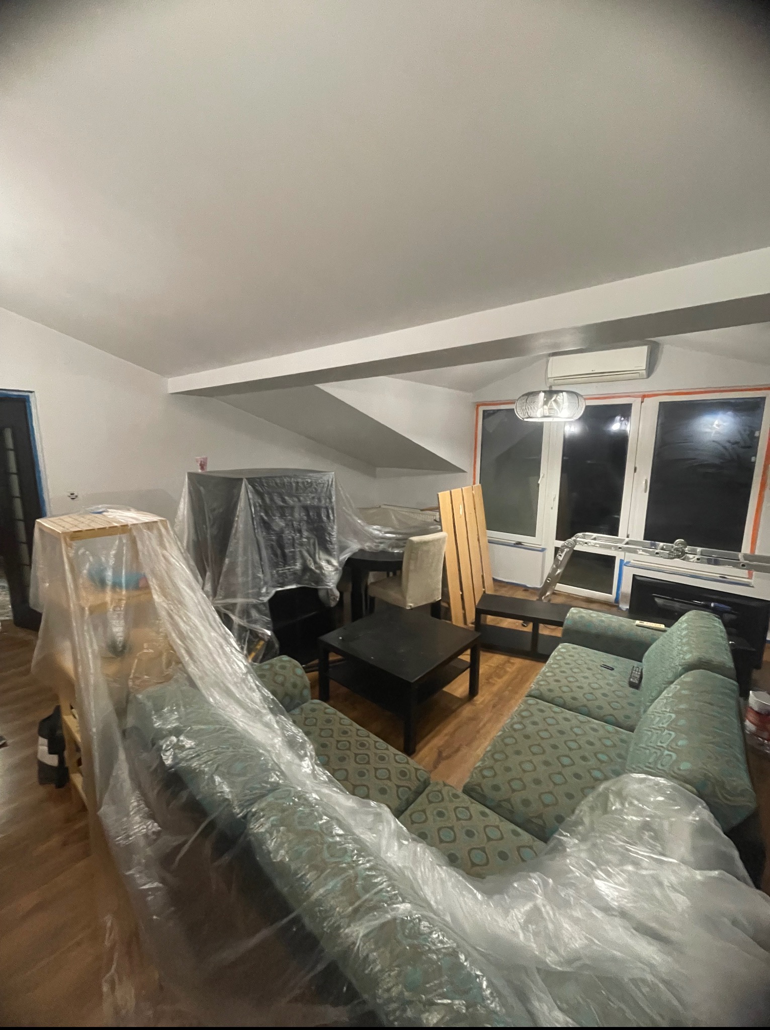Pokój zabezpieczony folią malarską przed remontem. Meble i sprzęty zakryte, widoczne okna i drabina. Przygotowania do prac remontowych.