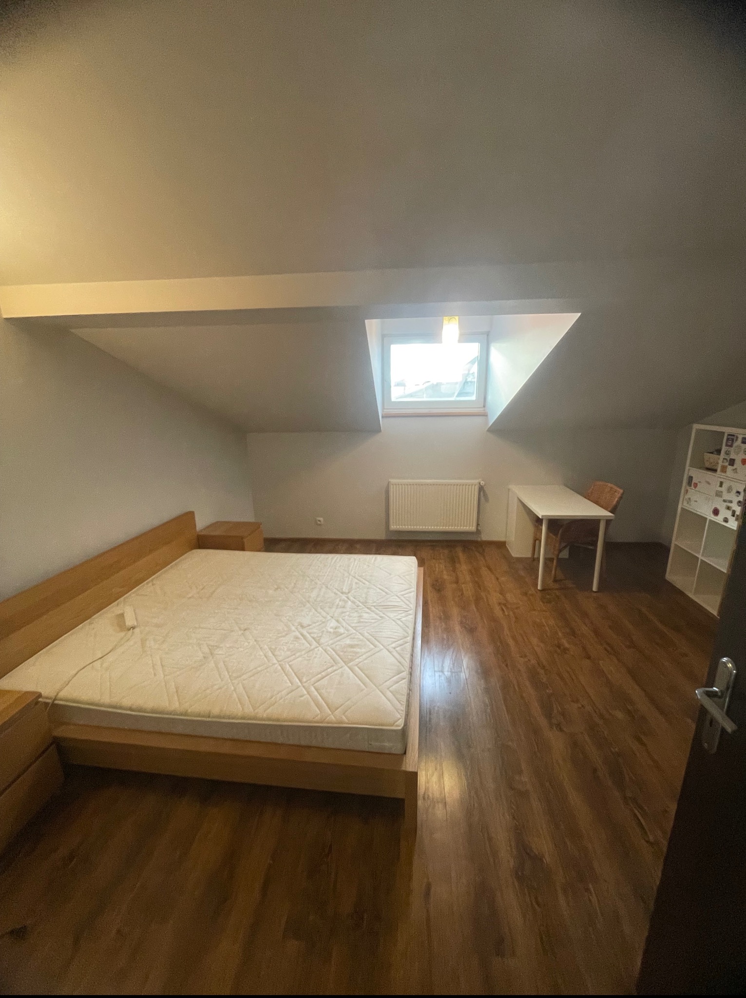 Pokój na poddaszu z drewnianą podłogą, łóżkiem i oknem dachowym. Ściany pomalowane na jasny kolor. Widoczne biurko, krzesło i regał.