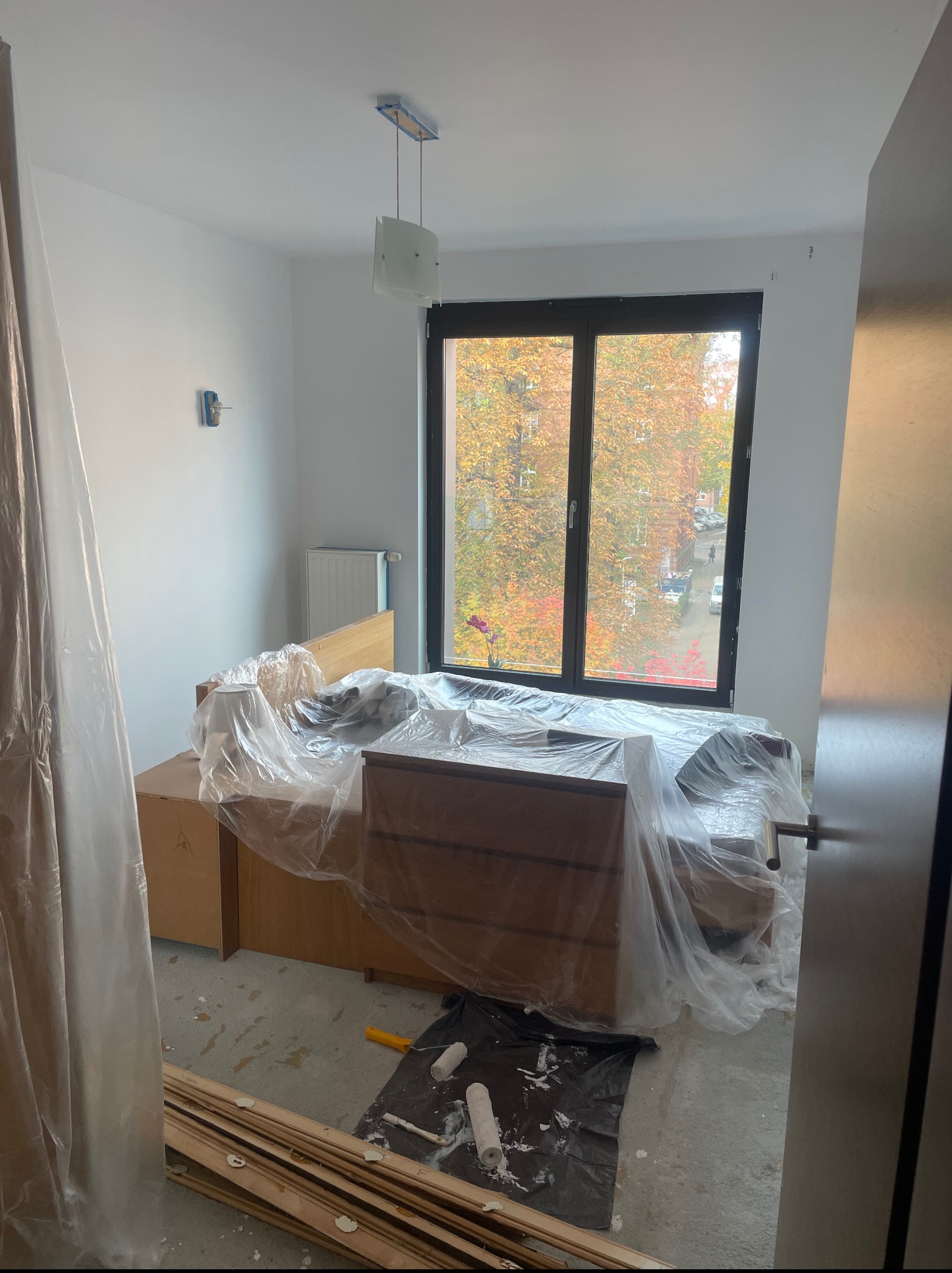 Pokój w trakcie malowania: meble zabezpieczone folią, wałki malarskie na podłodze, widok na jesienne drzewa za oknem. Świeżo pomalowane ściany w jasnym odcieniu.