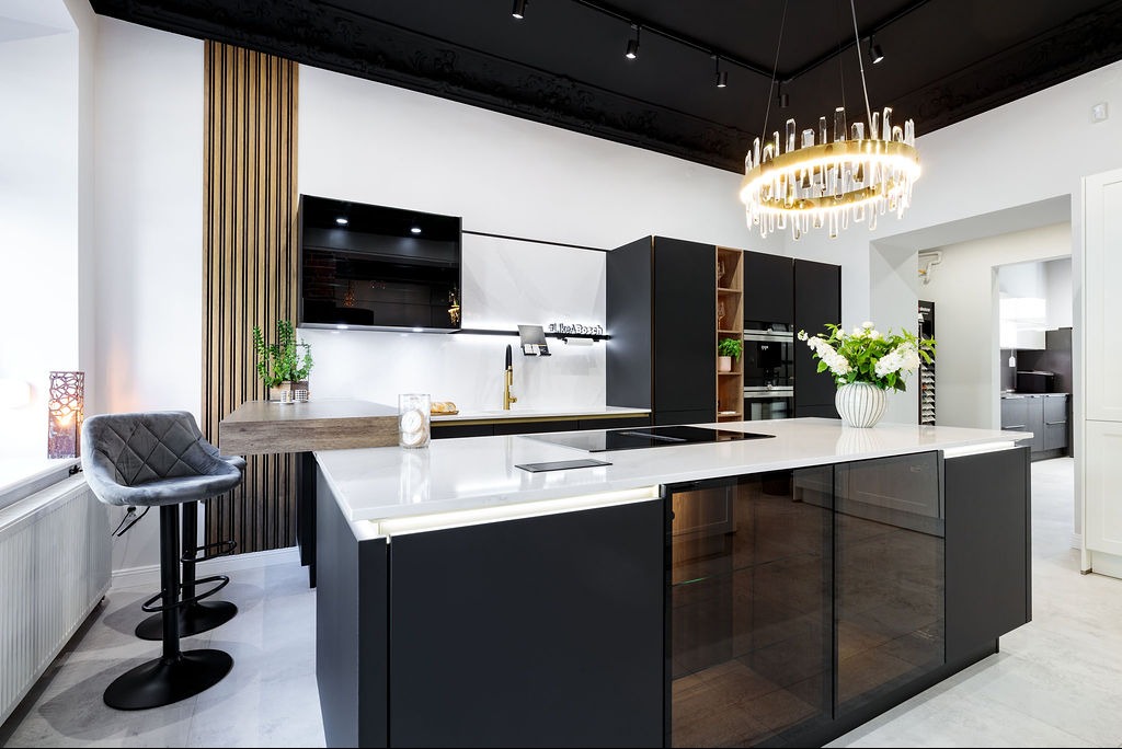 Nowoczesna kuchnia z wyspą, czarnymi szafkami i złotymi akcentami. Oświetlenie LED podkreśla marmurowy blat. Krzesło barowe i drewniane lamele dodają elegancji.