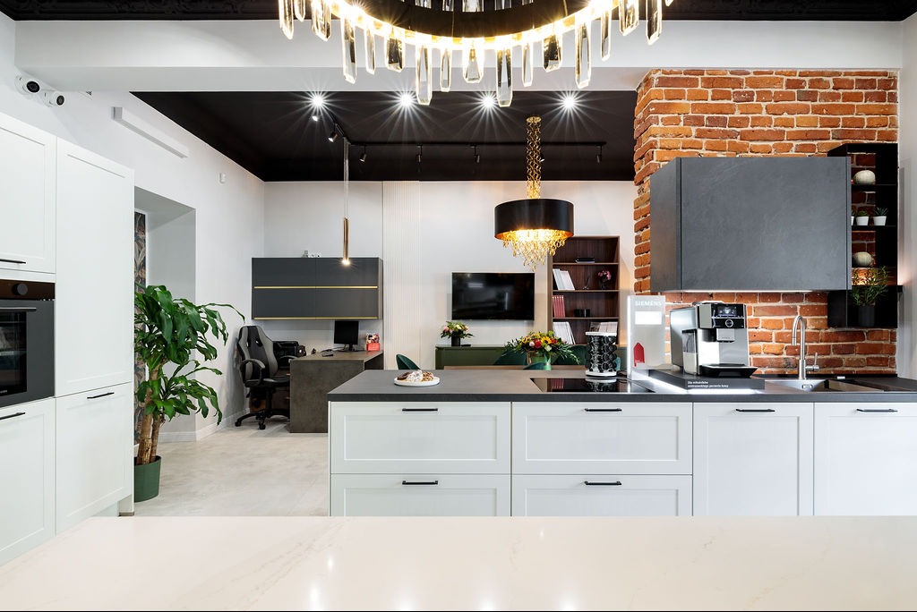 Nowoczesna kuchnia z białymi szafkami, czarnym blatem i ceglaną ścianą. W tle widoczne biurko i krzesło gamingowe. Nad blatem kryształowy żyrandol.