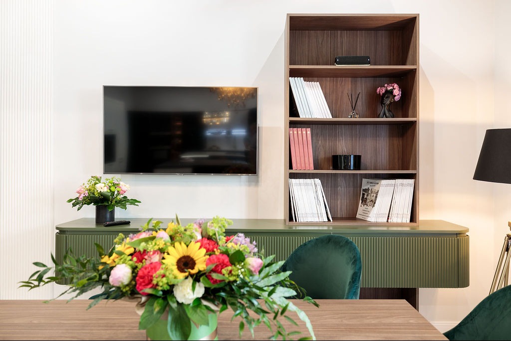 Nowoczesny salon: TV na ścianie, zielona szafka RTV z ryflowanym frontem, drewniany regał i bukiet kwiatów na stole. Wnętrze w stonowanej kolorystyce.