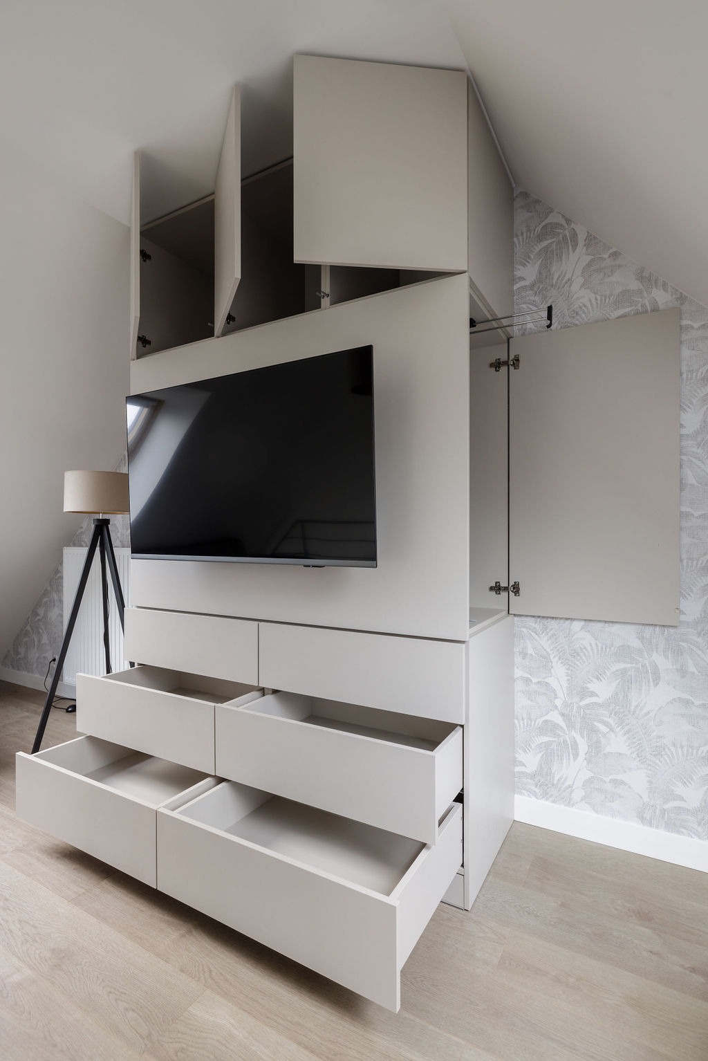 Biała zabudowa meblowa pod skosem ze schowkami, szafkami i szufladami. Zamontowany telewizor. Otwarta szafka i szuflady eksponują wnętrze. Lampa na trójnogu w tle.