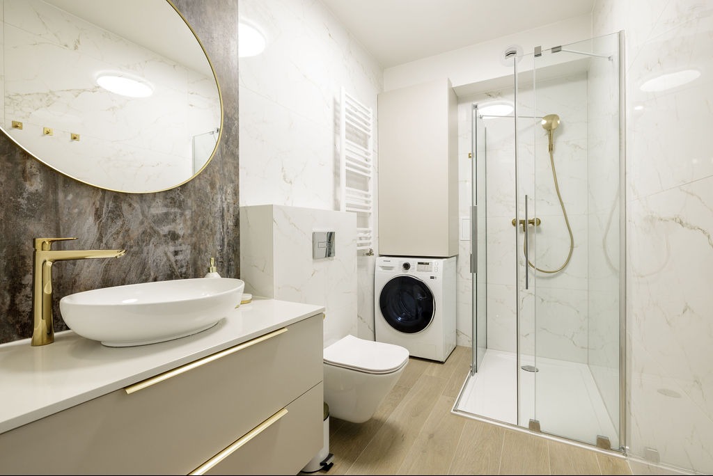 Jasna łazienka z marmurowymi płytkami, złotą armaturą i okrągłym lustrem. Kabina prysznicowa ze złotym zestawem, pralka i toaleta w tle.