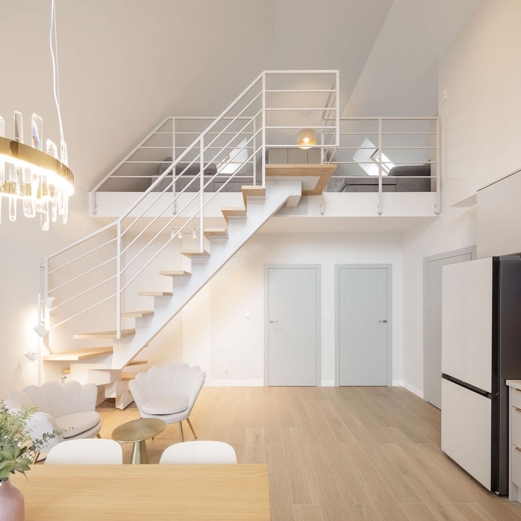 Jasne, przestronne wnętrze z antresolą i schodami o białej balustradzie. Widoczne krzesła muszelki, drewniana podłoga i nowoczesna lampa wisząca.