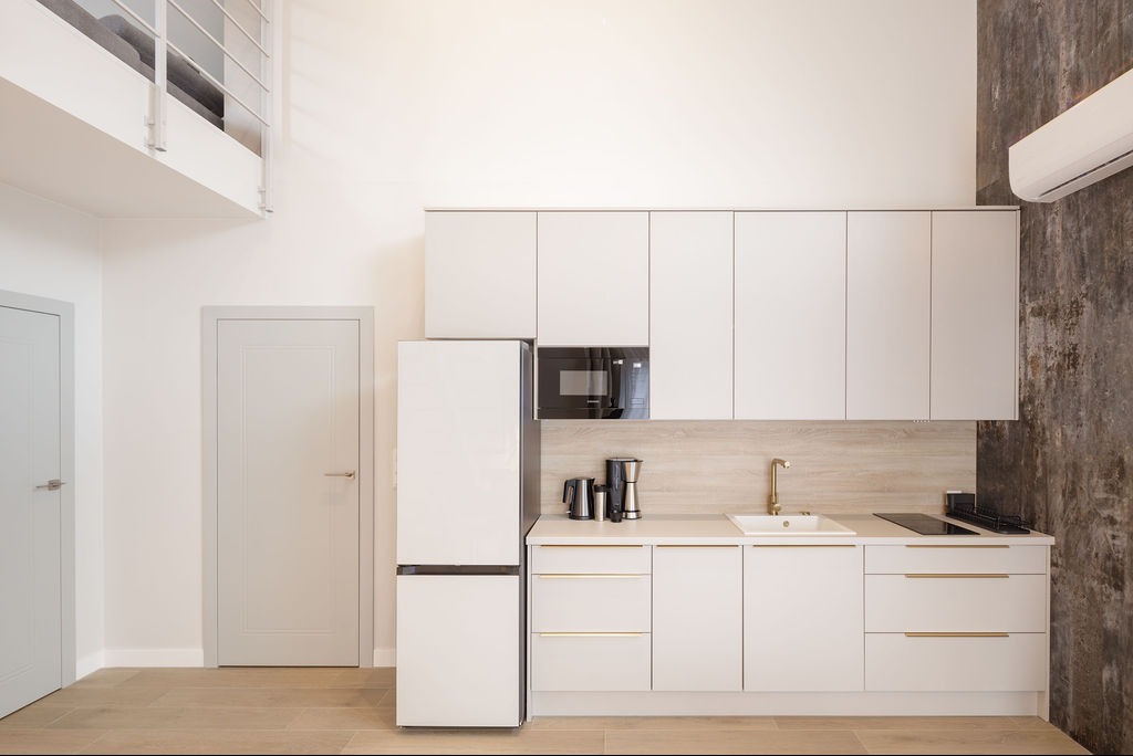 Jasna kuchnia z białymi szafkami i złotymi akcentami, widok na antresolę. Nowoczesny design z elementami loftu, oświetlenie punktowe i minimalistyczny styl.