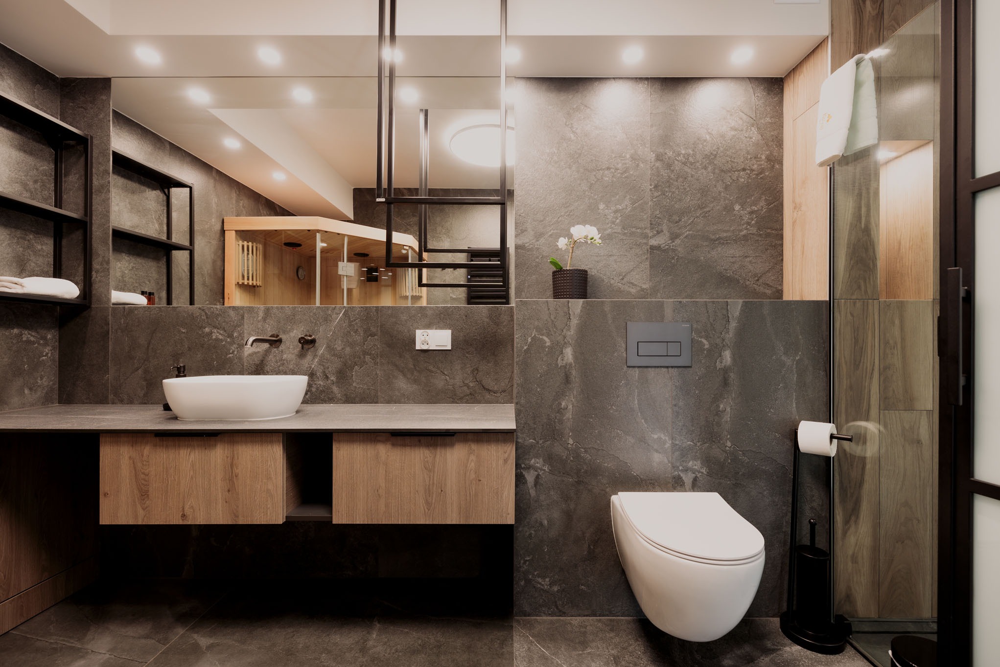 Nowoczesna łazienka z szarymi płytkami imitującymi kamień, drewnianą szafką pod umywalką i sauną widoczną w lustrze. Minimalistyczny design z akcentami drewna.