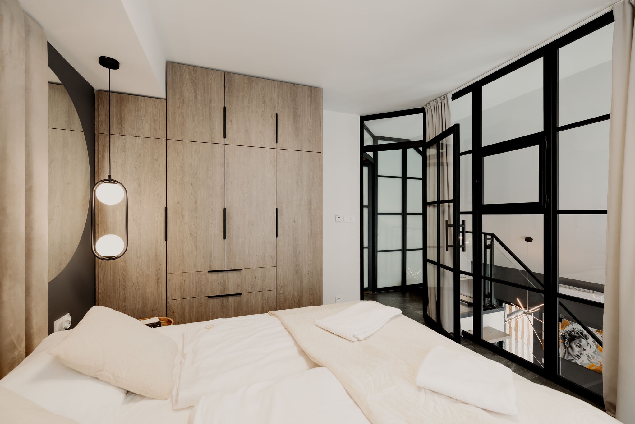 Sypialnia z zabudową szaf z jasnego drewna i przeszklonymi drzwiami w stylu loft. Oświetlenie punktowe i tekstylia w odcieniach bieli tworzą przytulny klimat.