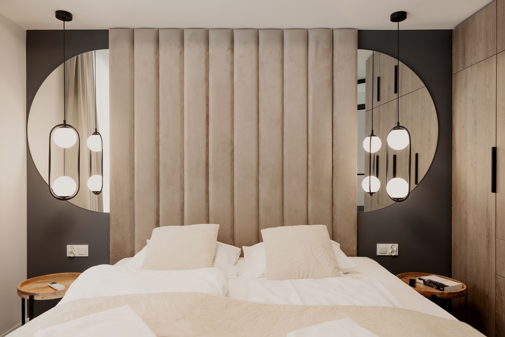 Elegancka sypialnia w neutralnych barwach: beżowe panele ścienne za łóżkiem, okrągłe lustra z nowoczesnymi lampami wiszącymi i drewniana szafa w tle.