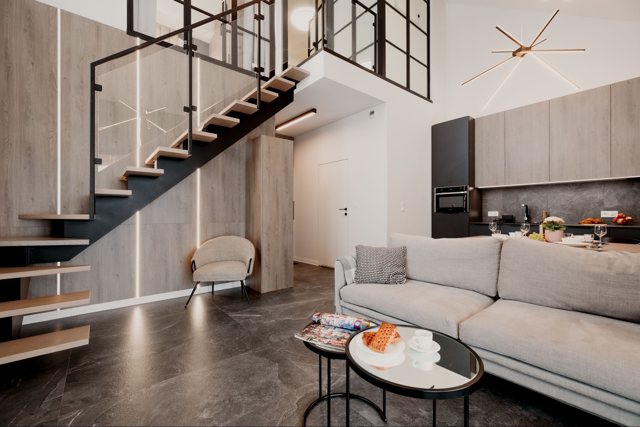 Nowoczesne wnętrze z drewnianymi schodami i szklaną balustradą. Minimalistyczny design z elementami drewna i czerni. Widoczny fragment kuchni i salonu.