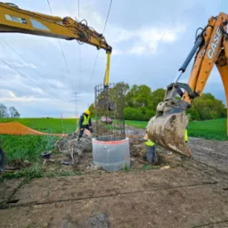 Montaż studni kręgowej z użyciem ciężkiego sprzętu na tle zielonego pola i pochmurnego nieba. Widoczne linie wysokiego napięcia.