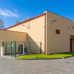Energetyka i Budownictwo Inwestycje Sp. z o.o. - Zewnętrzny widok stacji transformatorowej z zielonym ogrodzeniem i szarym brukiem. Żółty budynek z czerwonymi elementami, oświetlenie zewnętrzne i drabina na dachu.