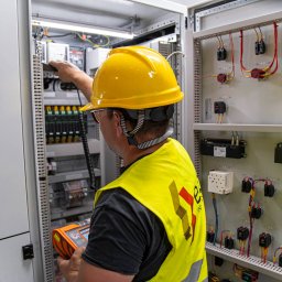Energetyka i Budownictwo Inwestycje Sp. z o.o. - Elektryk w kasku i kamizelce mierzy parametry w szafie sterowniczej. Widoczne przewody, zabezpieczenia i aparatura kontrolno-pomiarowa w rozdzielni elektrycznej.