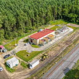 Energetyka i Budownictwo Inwestycje Sp. z o.o. - Widok z lotu ptaka na stację transformatorową z czerwonym dachem, otoczoną zielenią i lasem, z linią kolejową w tle. Na terenie stacji zaparkowane samochody i sprzęt budowlany.