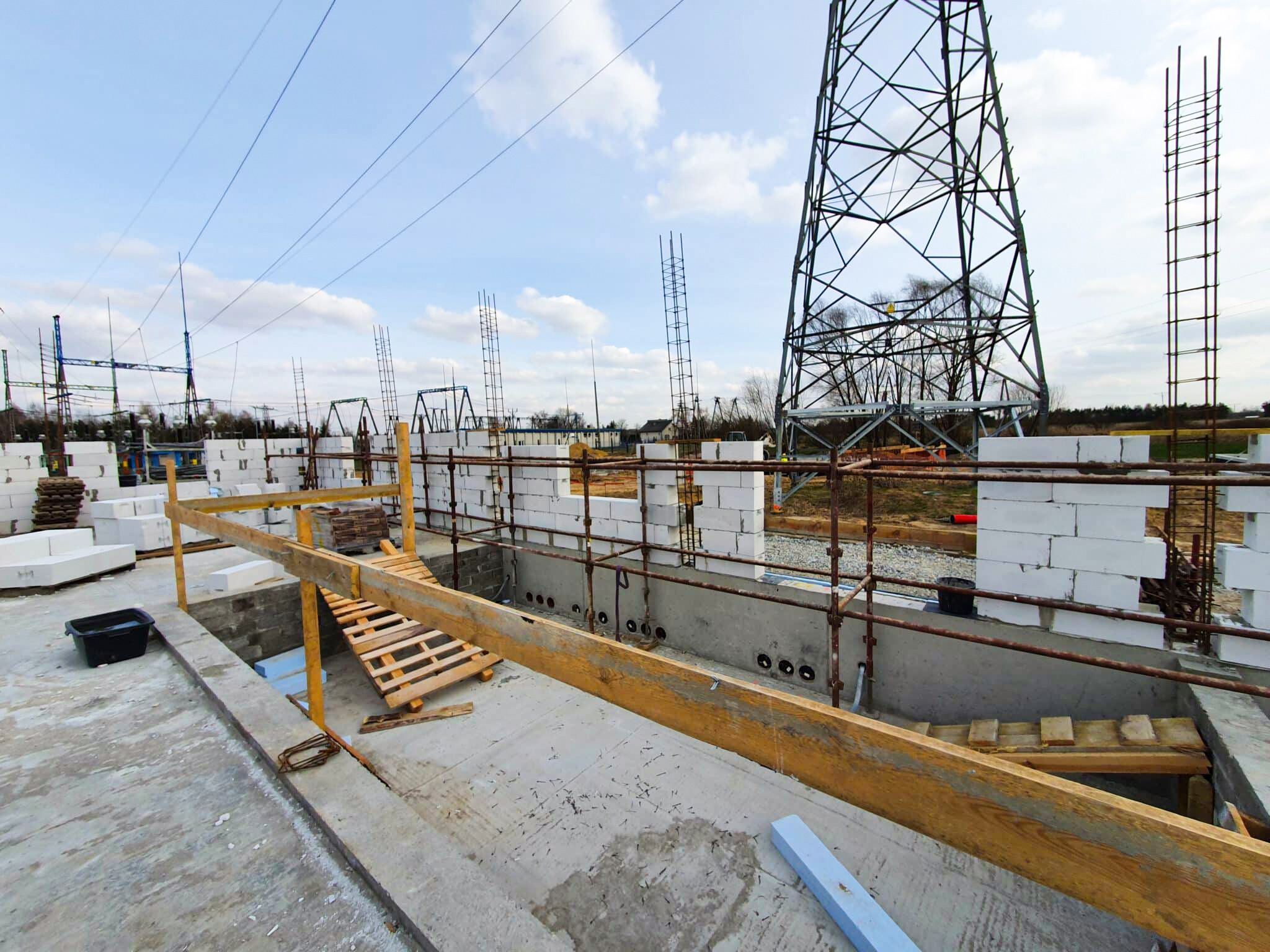 Budowa fundamentów z bloczków betonu komórkowego i widoczną konstrukcją wsporczą linii wysokiego napięcia w tle, w trakcie realizacji.