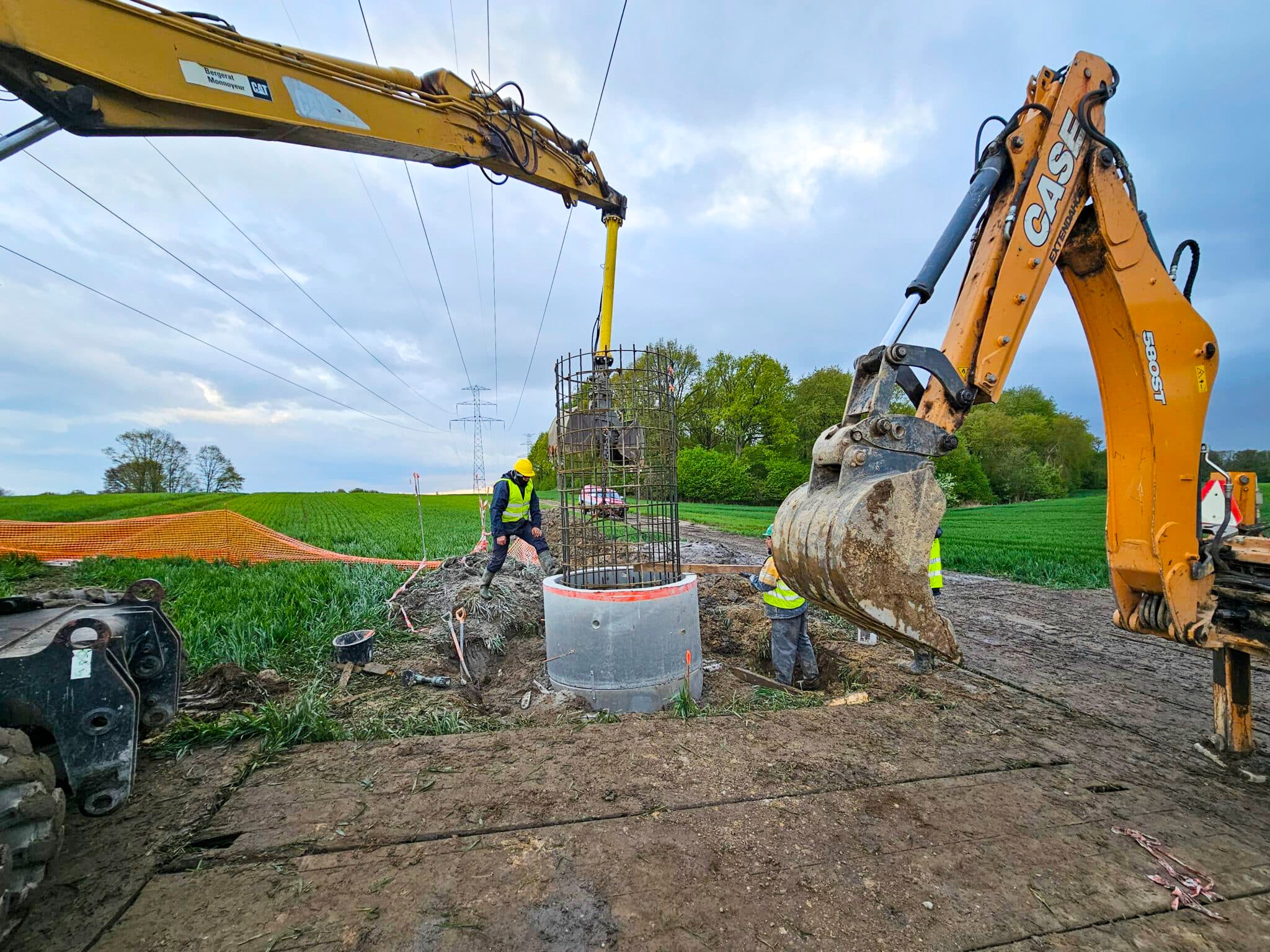 Montaż studni kręgowej z użyciem ciężkiego sprzętu na tle zielonego pola i pochmurnego nieba. Widoczne linie wysokiego napięcia.