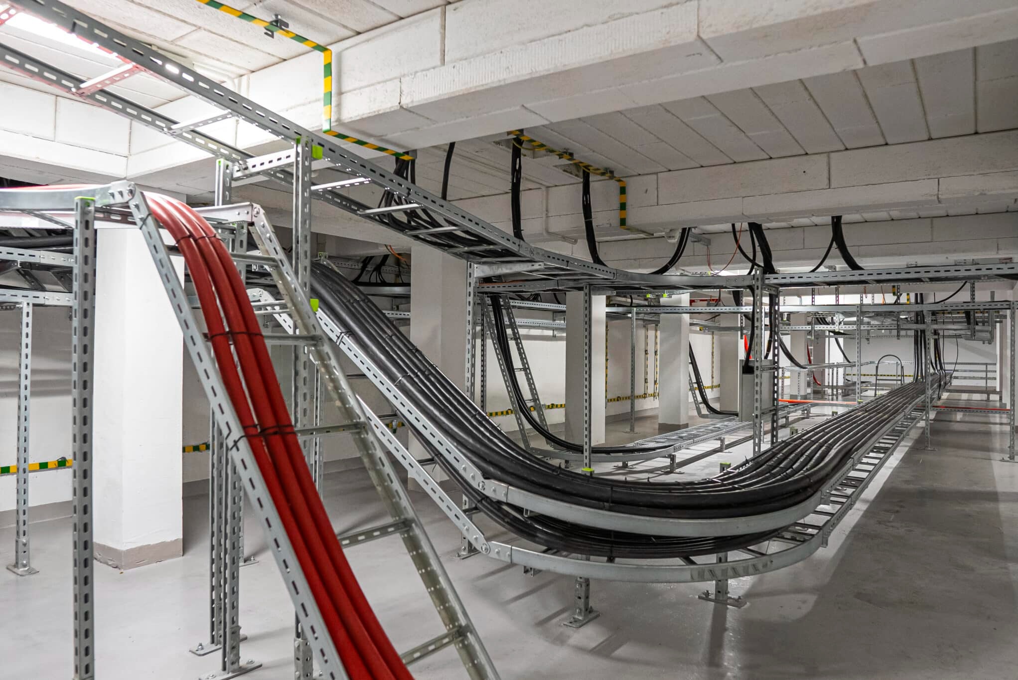 Profesjonalny montaż tras kablowych z przewodami wewnątrz budynku. Czerwone i czarne kable ułożone na metalowych konstrukcjach pod stropem.